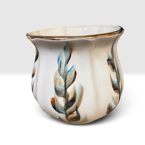 May include: A white ceramic planter with a scalloped rim and a gold-painted edge. The exterior features hand-painted vertical leaf motifs in shades of blue, brown, and black, arranged in a repeating pattern around the curved, fluted body.