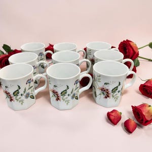 May include: A set of white ceramic mugs with a floral pattern featuring red, pink, blue, and green flowers and leaves. The mugs have a hexagonal shape and a curved handle. Red roses are arranged around the mugs.