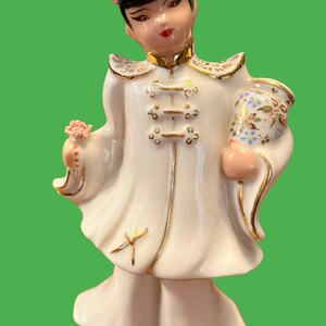 May include: A porcelain figurine of a person wearing a white robe with gold accents and a black hat. The figurine is holding a pink flower in one hand and a small, decorated vase in the other.