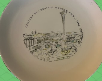 May include: A white plate featuring a detailed illustration of the Seattle World's Fair 1962, including the Space Needle and surrounding buildings. The text "Century 21 Seattle World's Fair 1962" is printed around the image.