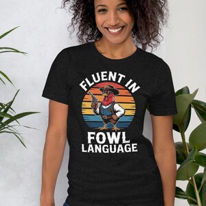 Può includere: T-shirt grigio scuro con un gallo dei cartoni animati con cappello da cowboy e gilet, che tiene una pistola. La maglietta presenta la scritta "FLUENT IN FOWL LANGUAGE" in un design circolare con uno sfondo di tramonto. La modella indossa jeans blu.