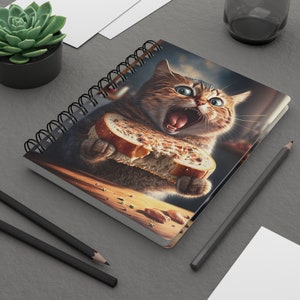 May include: Spiral-bound notebook featuring a digitally painted image of a tabby cat with wide eyes, mid-bite on a slice of bread. The cat's mouth is open, and the bread is partially eaten. The notebook has a black spiral binding.