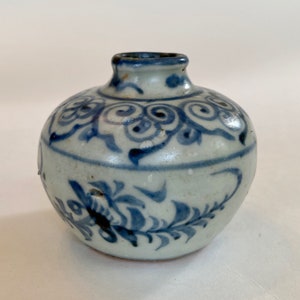Peut inclure: Un petit vase rond en céramique blanche avec des motifs floraux bleus et blancs. Le vase a une ouverture étroite en haut.