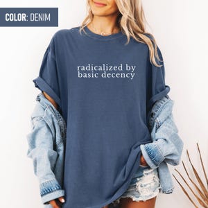 Op de afbeelding: Een denimblauw T-shirt met de tekst "radicalized by basic decency" in wit gedrukt.