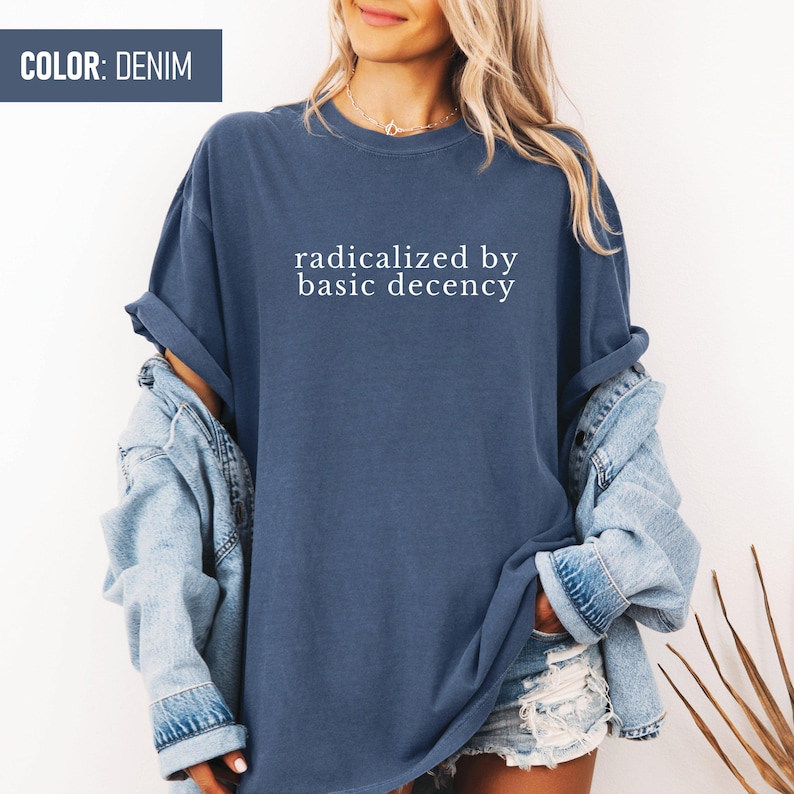 Op de afbeelding: Een denimblauw T-shirt met de tekst "radicalized by basic decency" in wit gedrukt.