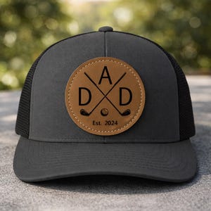 May include: A gray and black trucker hat with a brown leather patch. The patch features the word "DAD" with crossed golf clubs and a golf ball, and the text "Est. 2024". The hat has a curved brim and a mesh back.