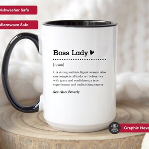 May include: A white ceramic mug with a black handle and interior, featuring the black printed text Boss Lady, noun, and a definition describing a strong and intelligent person who completes tasks with grace and confidence, including the text See Also: Beverly.