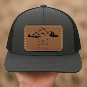 May include: Black trucker hat with a brown leather patch featuring a mountain silhouette, two arrows crossed with the letters E & S, the date 10.12.24, and the text "Bass Lake, CA".