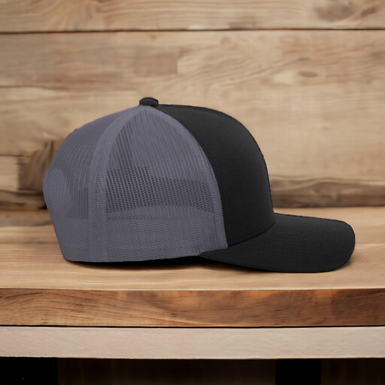 May include: Black and gray trucker hat with a curved brim. The front panels and brim are black, while the back mesh panel is gray. The hat is sitting on a wooden surface.