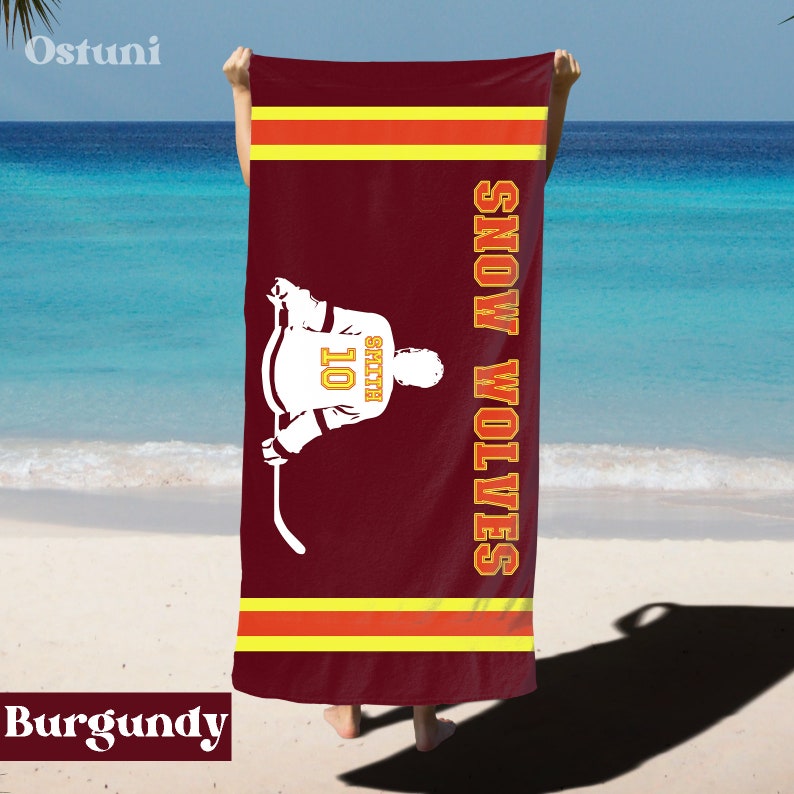 May include: Burgundy beach towel with a hockey player silhouette and the text "SNOW WOLVES" in white. The player is wearing a jersey with the number "10" and the name "SMITH" on the back.