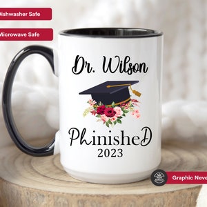 May include: White ceramic mug with a black rim and handle. The mug has a black graphic of a graduation cap with flowers and the text "Dr. Wilson Phinished 2023".