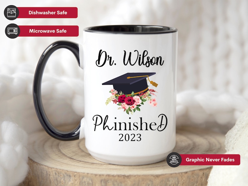 May include: White ceramic mug with a black rim and handle. The mug has a black graphic of a graduation cap with flowers and the text "Dr. Wilson Phinished 2023".