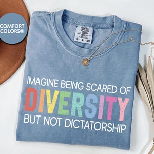 May include: A blue Comfort Colors t-shirt with the text "IMAGINE BEING SCARED OF DIVERSITY BUT NOT DICTATORSHIP" in colorful letters. A gold necklace with a pendant is draped across the shirt.