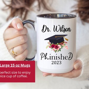 May include: White ceramic mug with a black rim and handle. The mug has a black and white graphic of a graduation cap with flowers and the text "Dr. Wilson Phinished 2023".