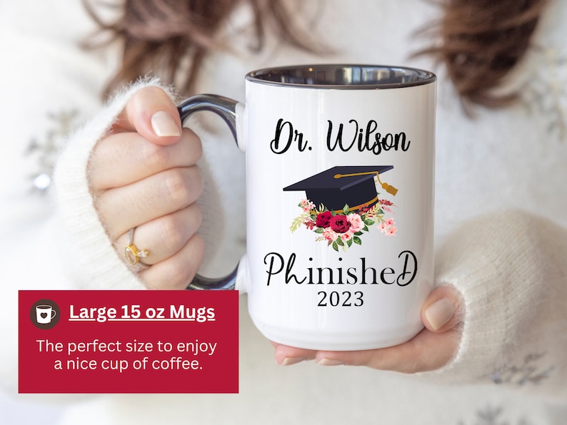 May include: White ceramic mug with a black rim and handle. The mug has a black and white graphic of a graduation cap with flowers and the text "Dr. Wilson Phinished 2023".