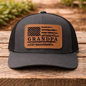 May include: A gray trucker hat with a black mesh back and a curved brim. The hat features a brown leather patch with an American flag design and the word "GRANDPA" in bold letters. The patch also includes names.