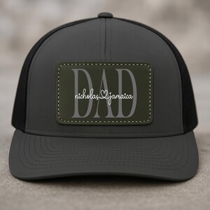 May include: A dark gray trucker hat with a black mesh back. The hat features a rectangular patch with the word "DAD" in large gray letters, and the names "nicholas & jamaica" in white script.