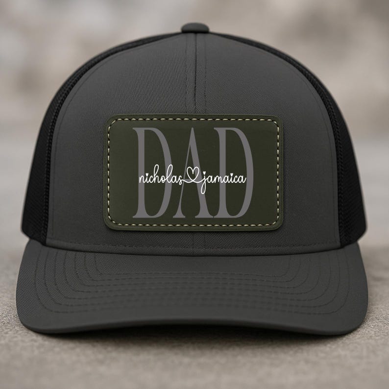 May include: A dark gray trucker hat with a black mesh back. The hat features a rectangular patch with the word "DAD" in large gray letters, and the names "nicholas & jamaica" in white script.