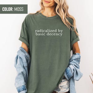 Op de afbeelding: Een mosgroen T-shirt met de tekst "radicalized by basic decency" in wit gedrukt op de voorkant.