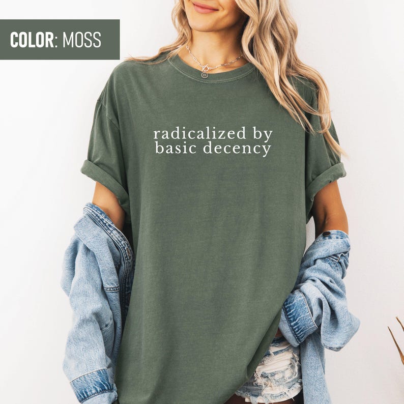 Op de afbeelding: Een mosgroen T-shirt met de tekst "radicalized by basic decency" in wit gedrukt op de voorkant.