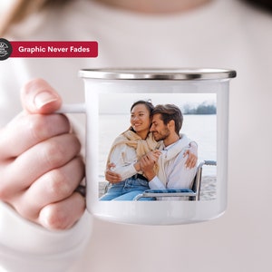 K&ouml;nnte beinhalten: Eine wei&szlig;e Keramiktasse mit einem Foto eines Paares, das sich an einem Strand umarmt. Die Tasse hat einen silbernen Rand und einen Henkel. Der Text "Graphic Never Fades" ist auf einem roten Banner &uuml;ber der Tasse gedruckt.