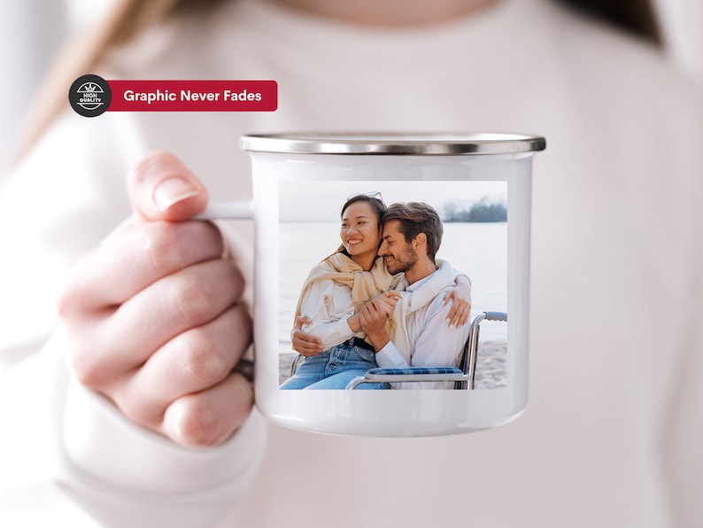 K&ouml;nnte beinhalten: Eine wei&szlig;e Keramiktasse mit einem Foto eines Paares, das sich an einem Strand umarmt. Die Tasse hat einen silbernen Rand und einen Henkel. Der Text "Graphic Never Fades" ist auf einem roten Banner &uuml;ber der Tasse gedruckt.