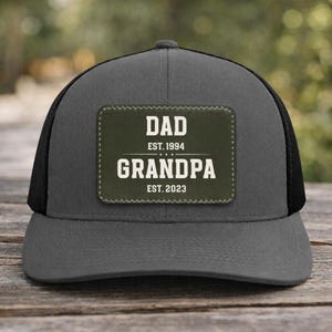 May include: A gray trucker hat with a black mesh back. The front features a rectangular patch with the words "DAD EST. 1994 GRANDPA EST. 2023" in white lettering. The hat is designed for everyday wear.