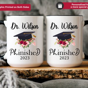 May include: Two white coffee mugs with black handles and black rims. The mugs have a graduation cap with flowers and the text "Dr. Wilson Phinished 2023" printed on them.