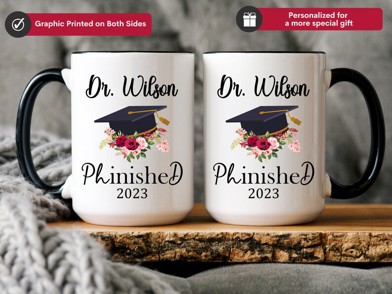 May include: Two white coffee mugs with black handles and black rims. The mugs have a graduation cap with flowers and the text "Dr. Wilson Phinished 2023" printed on them.