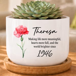 May include: White ceramic planter with a succulent plant. The planter features a pink flower illustration and the name "Theresa" with the text "Making life more meaningful, hearts more full, and the world brighter since 1946."