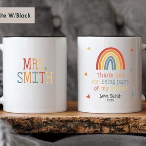 May include: Two white ceramic mugs with black rims. One mug has the text "MRS. SMITH" in colorful letters. The other mug has a rainbow design and the text "Thank you for being part of my story! Love, Sarah 2025".