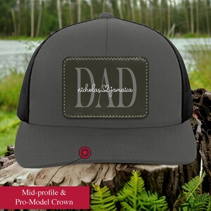 May include: A charcoal gray trucker hat with a black mesh back. The hat features a rectangular patch with the word "DAD" and the names "nicholas" and "jamaica" in white script. The hat has a mid-profile and pro-model crown.
