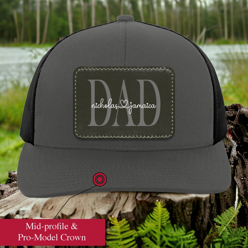 May include: A charcoal gray trucker hat with a black mesh back. The hat features a rectangular patch with the word "DAD" and the names "nicholas" and "jamaica" in white script. The hat has a mid-profile and pro-model crown.