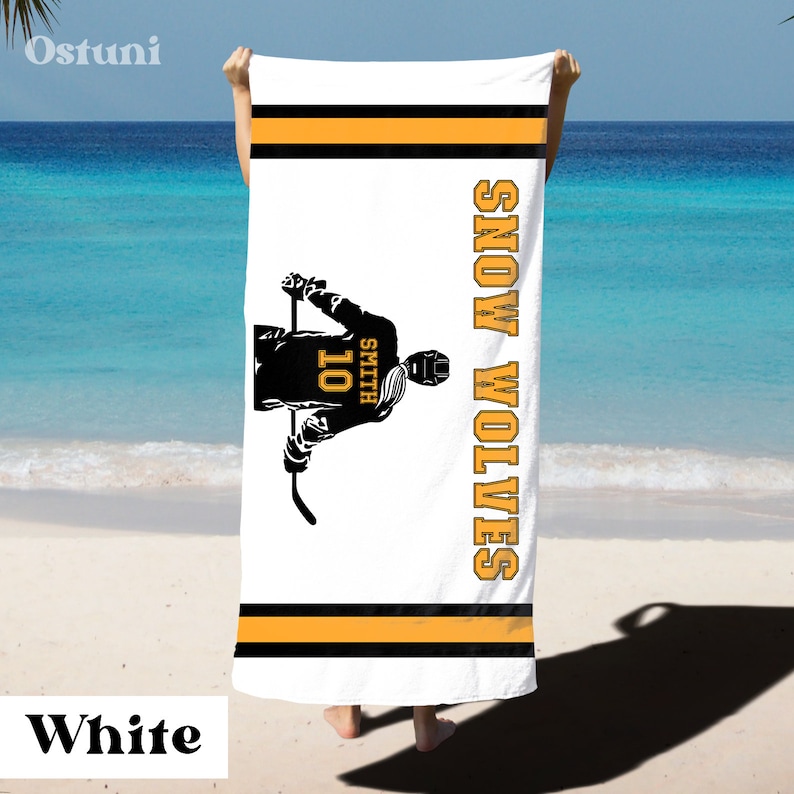 May include: White beach towel with a black silhouette of a hockey player holding a stick. The player is wearing a jersey with the number 10 and the name Smith. The text "SNOW WOLVES" is printed vertically on the right side of the towel. The towel has black and orange stripes.