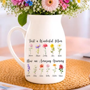May include: A white ceramic pitcher featuring a handle and a printed design of two rows of colorful watercolor flowers. The text reads First a Wonderful Mom and Now an Amazing Grammy, with individual names printed below each flower illustration.