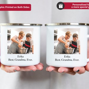 K&ouml;nnte beinhalten: Zwei wei&szlig;e Emailletassen mit einem Foto einer Gro&szlig;mutter und eines Enkels beim Backen. Die Tassen haben einen silbernen Rand und einen Henkel. Der Text "Erika Best. Grandma. Ever." ist auf den Tassen aufgedruckt.