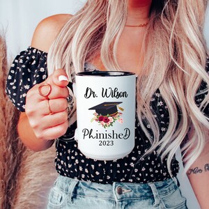 May include: White coffee mug with a black rim and handle. The mug has a black graduation cap with a red and white floral design and the text "Dr. Wilson Phinished 2023".