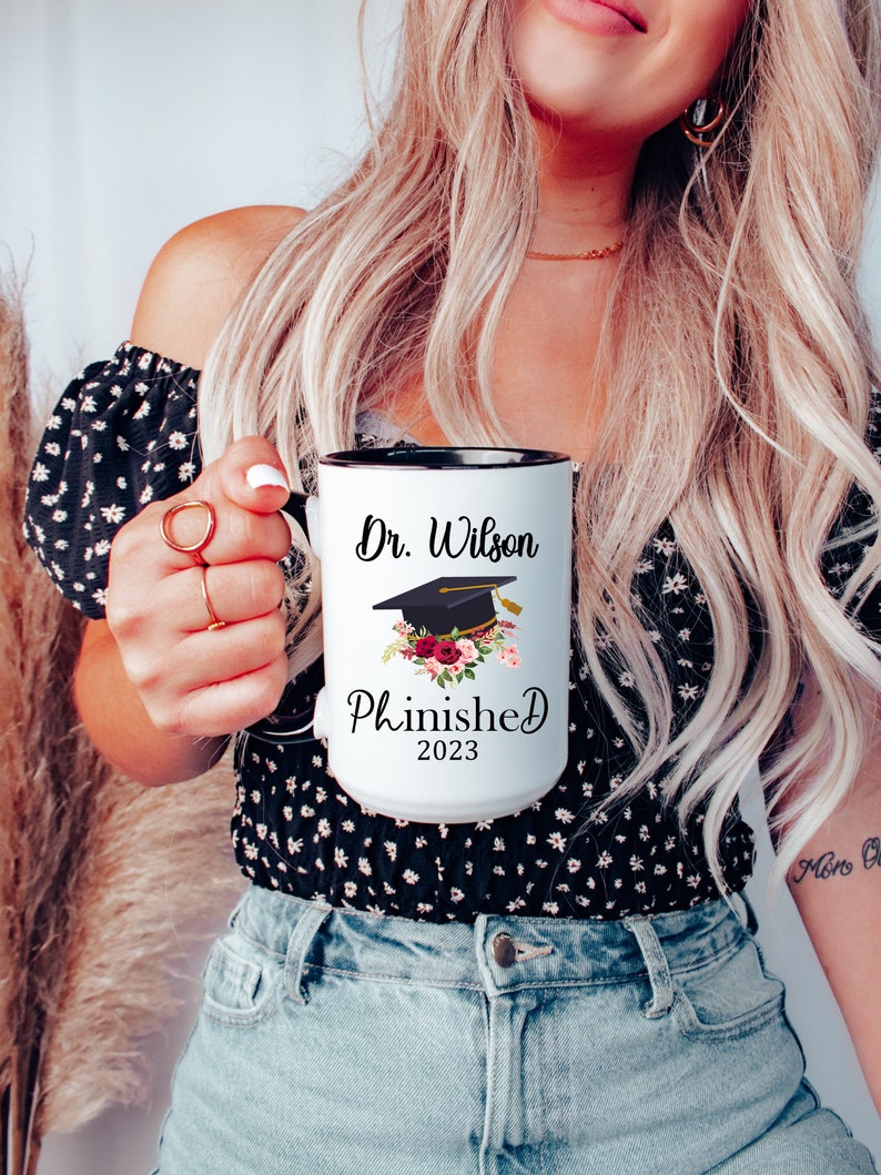 May include: White coffee mug with a black rim and handle. The mug has a black graduation cap with a red and white floral design and the text "Dr. Wilson Phinished 2023".