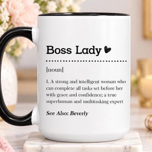 May include: A white ceramic mug with a black handle and rim, featuring the black printed text Boss Lady, followed by the definition of a strong and intelligent person who completes tasks with grace and confidence, ending with See Also: Beverly.