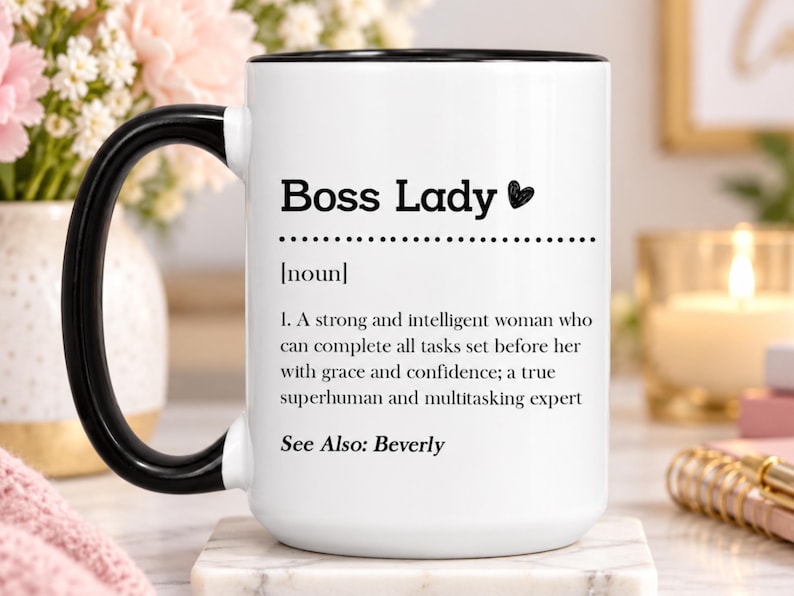 May include: A white ceramic mug with a black handle and rim, featuring the black printed text Boss Lady, followed by the definition of a strong and intelligent person who completes tasks with grace and confidence, ending with See Also: Beverly.