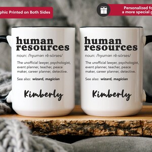 May include: Two white ceramic mugs with black handles. The mugs feature a black graphic that reads "human resources" with a definition and the name "Kimberly" printed below. The mugs are resting on a wooden surface.