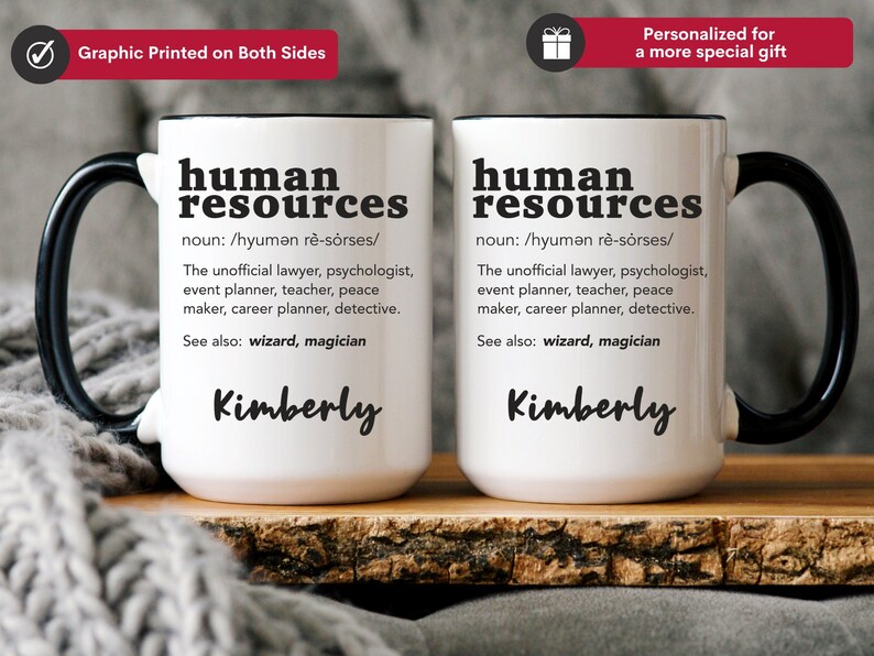 May include: Two white ceramic mugs with black handles. The mugs feature a black graphic that reads "human resources" with a definition and the name "Kimberly" printed below. The mugs are resting on a wooden surface.