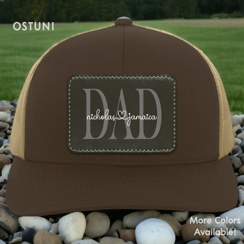 May include: A brown trucker hat with a tan mesh back. The front features a rectangular patch with the word "DAD" and the names "nicholas" and "jamaica" in white script. The hat is sitting on a bed of rocks.