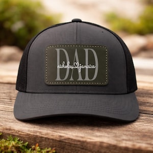 May include: A gray trucker hat with a black mesh back. The front features a rectangular patch with the word "DAD" and the names "nicholas jamaica" in white lettering. The hat is sitting on a wooden surface.