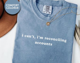 May include: A blue Comfort Colors t-shirt with the text "i can't, i'm reconciling accounts" in white. A gold necklace with a pendant is draped across the shirt. The shirt is folded on a white surface.