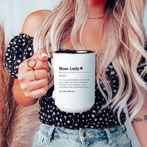 May include: A white ceramic mug with a black interior and handle, featuring the black printed text Boss Lady followed by a definition and the phrase See Also: Beverly. The mug has a cylindrical shape and a smooth, glossy finish.