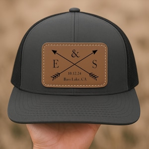 May include: Black trucker hat with a brown leather patch featuring two arrows pointing towards each other with the letters "E" and "S" on either side of an ampersand. The text "10.12.24 Bass Lake, CA" is printed below the arrows.