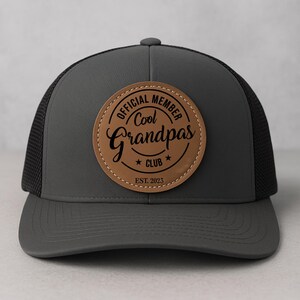 May include: A gray trucker hat with a black mesh back and a curved brim. The hat features a round, brown leather patch with the words "OFFICIAL MEMBER Cool Grandpas CLUB EST. 2023" in black lettering.