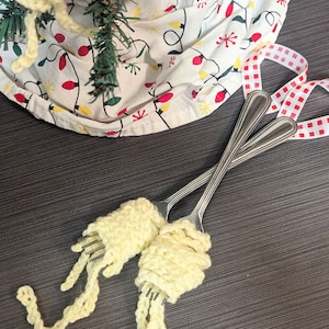 May include: Two crocheted yellow spaghetti noodle ornaments wrapped around silver forks. The ornaments are hanging from a Christmas tree.
