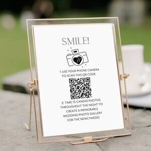 Può includere: Un cartello bianco con testo nero e un'icona di una macchina fotografica in bianco e nero. Il cartello dice "SMILE!" e chiede agli ospiti di usare la fotocamera del loro telefono per scansionare il codice QR per creare una galleria di foto di matrimonio.